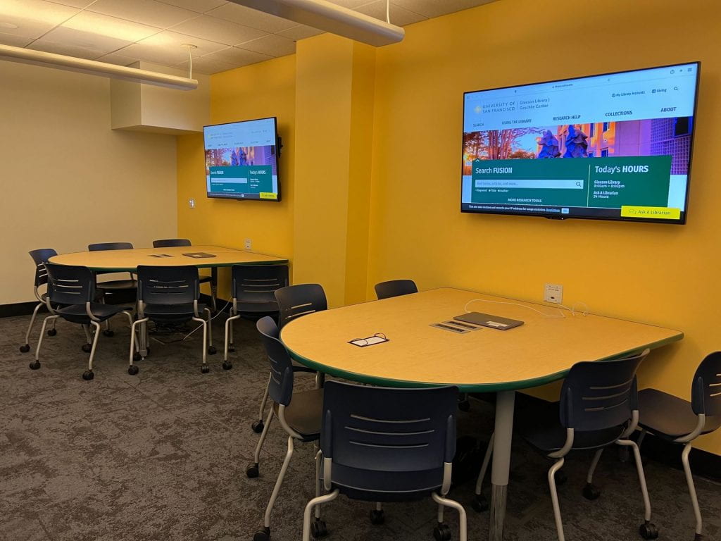 Two semi-round tables are set, flush against the wall. Above the tables are two screens which are displaying the library's homepage. Each table has 6 chairs around it. The tables are clean looking and each have a laptop on it.