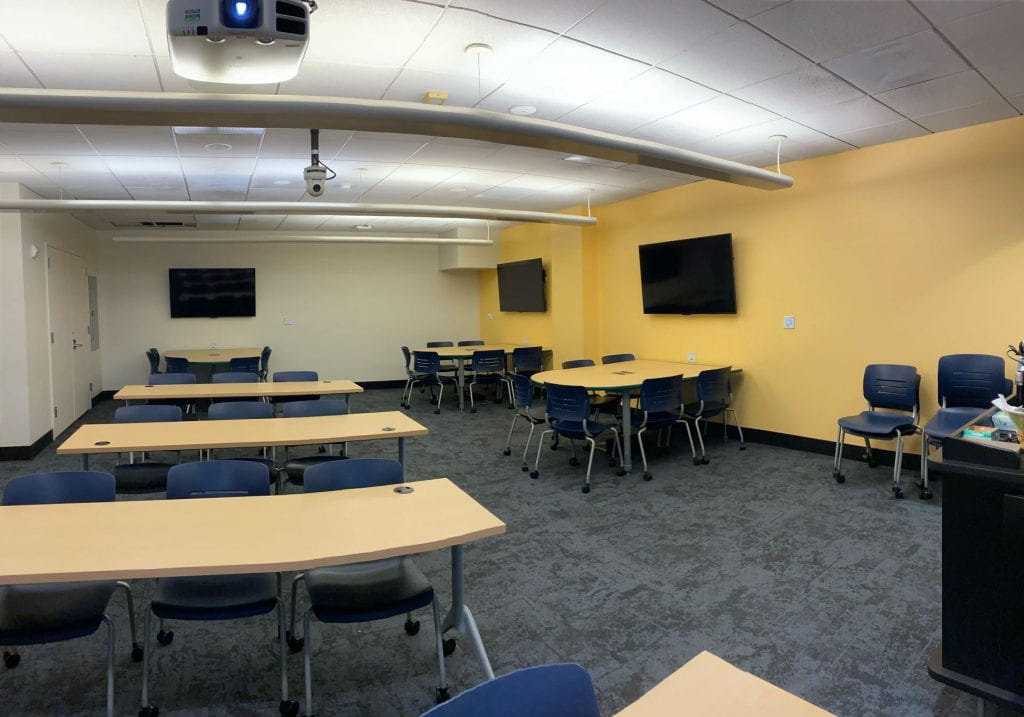 The view of the classroom from the front of the room where three rectangular tables are in a row in the center of the room. Along the perimeter of the room, there are three semi-round tables along with three screens. 