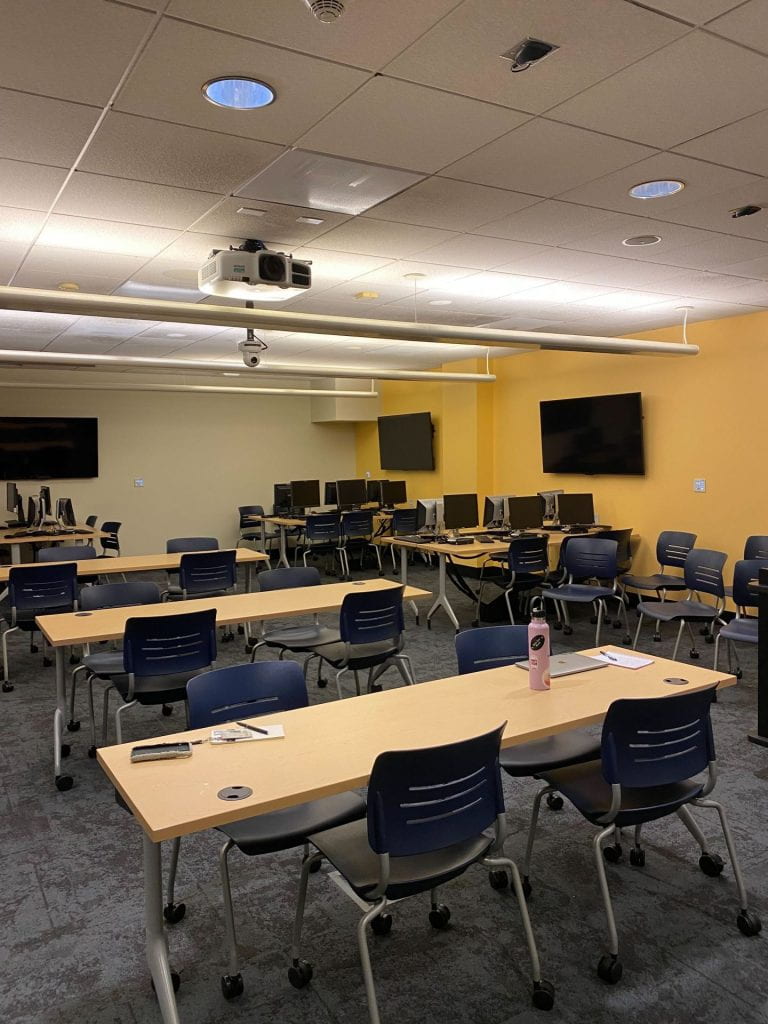 A previous configuration of the classroom which shows rectangular tables, and three stations with several PC desktops along the perimeter of the room.