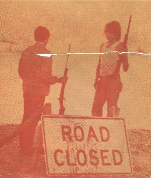 Two Native activists holding rifles stand guard behind Road Closed sign 