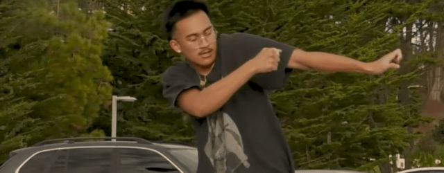 A dark-haired man in glasses and t-shirt dances in a parking lot against a backdrop of lush green trees.
