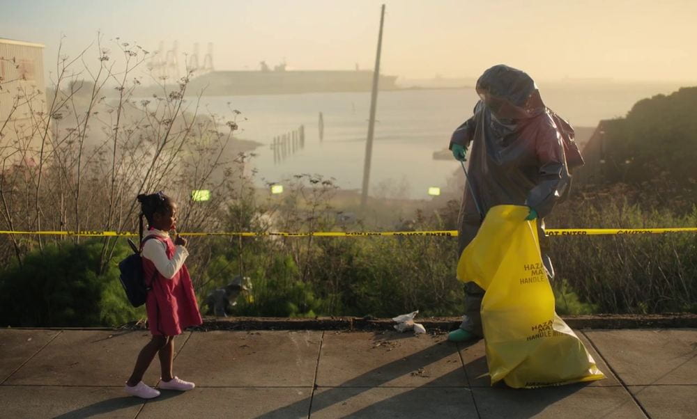 A man in a hazmat suit picks up trash while a young girl looks on